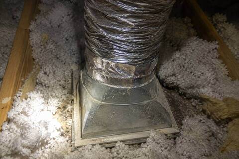 Flexible duct joining a sheet metal boot above a ceiling register, seen from the attic with insulation pulled back