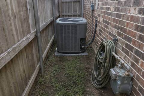 Outdoor condenser unit in a narrow side yard between a wooden fence and a brick wall, clearance tight on both sides