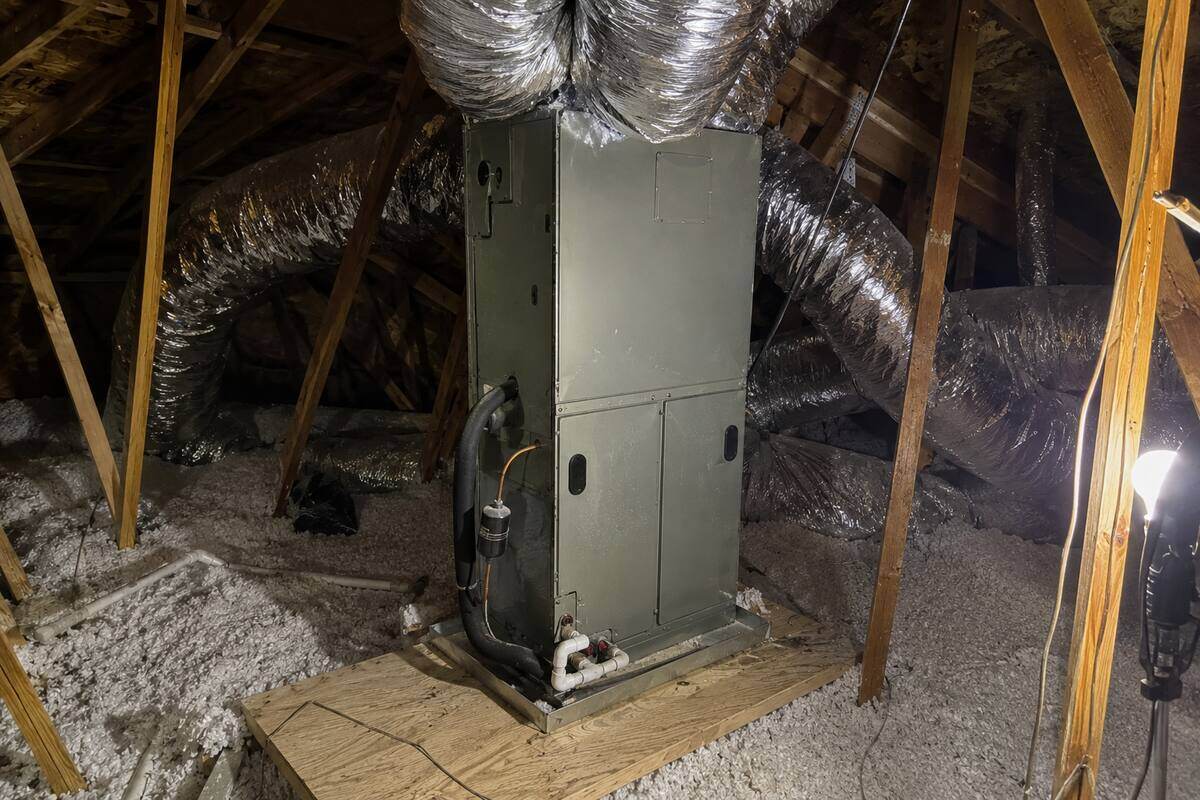 Air handler cabinet standing on a plywood platform in an attic, blown-in insulation and roof framing around it