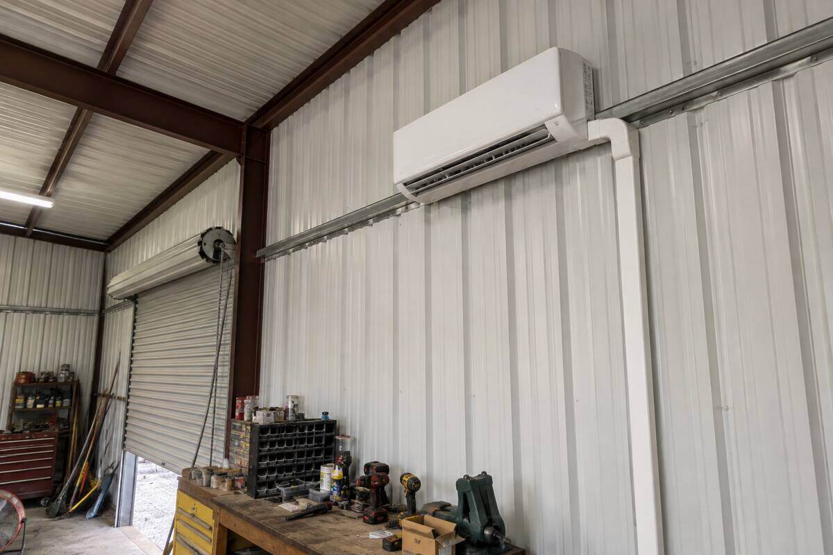 Wall-mounted ductless indoor unit high on the interior wall of a metal workshop, line set running down through the wall