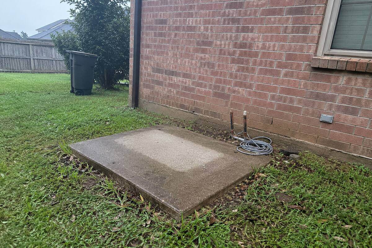 Empty concrete pad beside a brick wall with capped refrigerant line stubs standing where an outdoor unit was removed