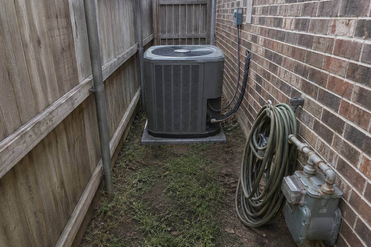 Outdoor condenser unit in a narrow side yard between a wooden fence and a brick wall, clearance tight on both sides